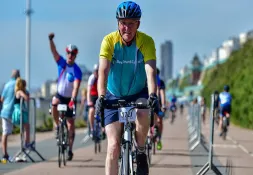 Man cycling in a charity bike ride for Bowel Cancer UK, wearing a helmet and branded jersey, with other cyclists and spectators along a coastal promenade.