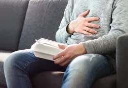 A man in a gray sweater sits on a couch holding a takeout container while placing a hand on his chest, suggesting difficulty eating or discomfort.