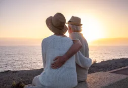 Older man and woman with their arms around each other sitting on a bench overlooking the sea watching the sunset