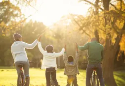 a mother, father and two children holding hands and riding bicycles on a field surrounded by trees 