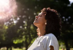 A young woman outdoors looking upward, with sunlight filtering through trees in the background. 