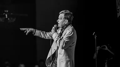 Black and white photo of a man speaking into a microphone on stage, pointing towards the audience with a spotlight highlighting him.