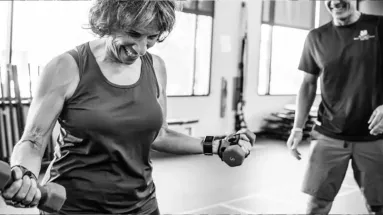 Black and white photo of a smiling woman lifting dumbbells in a fitness class, with an instructor in the background offering support.