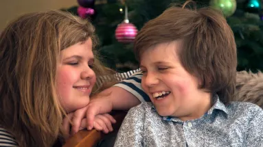 Two children smiling and laughing together in front of a decorated Christmas tree, sharing a joyful moment.