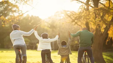 a mother, father and two children holding hands and riding bicycles on a field surrounded by trees 