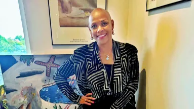 A woman (Sharon-Ann) smiling and wearing large silver earrings, a black and silver patterned cardigan, two silver necklaces and a black t-shirt. She has pink nail varnish on and is standing in front of a painting showing lightning, rain and a lake with houses along the side of it 