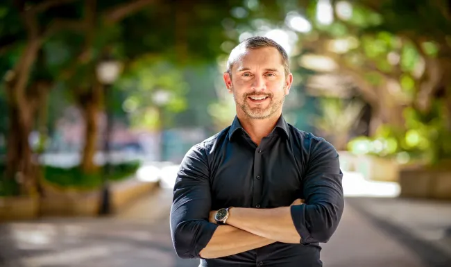 A man standing on a street with trees as the backdrop wearing a lack shirt with his arms crossed and sleeved rolled up